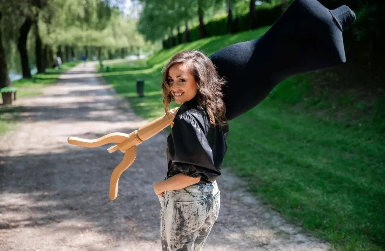 Nasmejana ženska hodi po gozdni poti in nosi črno krojaško lutko čez ramo medtem ko se ozira nazaj proti kameri