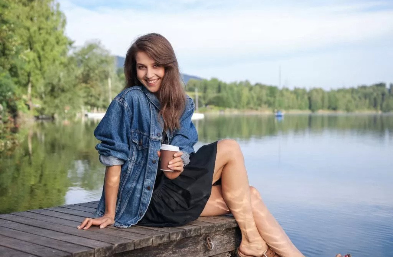 Nasmejana ženska sedi sproščeno na lesenem pomolu ob jezeru in v roki drži papirnato skodelico medtem ko jo obdaja mirna narava in odsev dreves v vodi