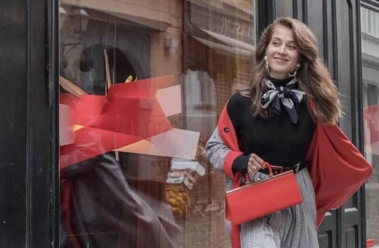 Elegantna ženska v črno rdeči obleki samozavestno hodi mimo izložbe in z nasmeškom uživa v mestnem sprehodu 