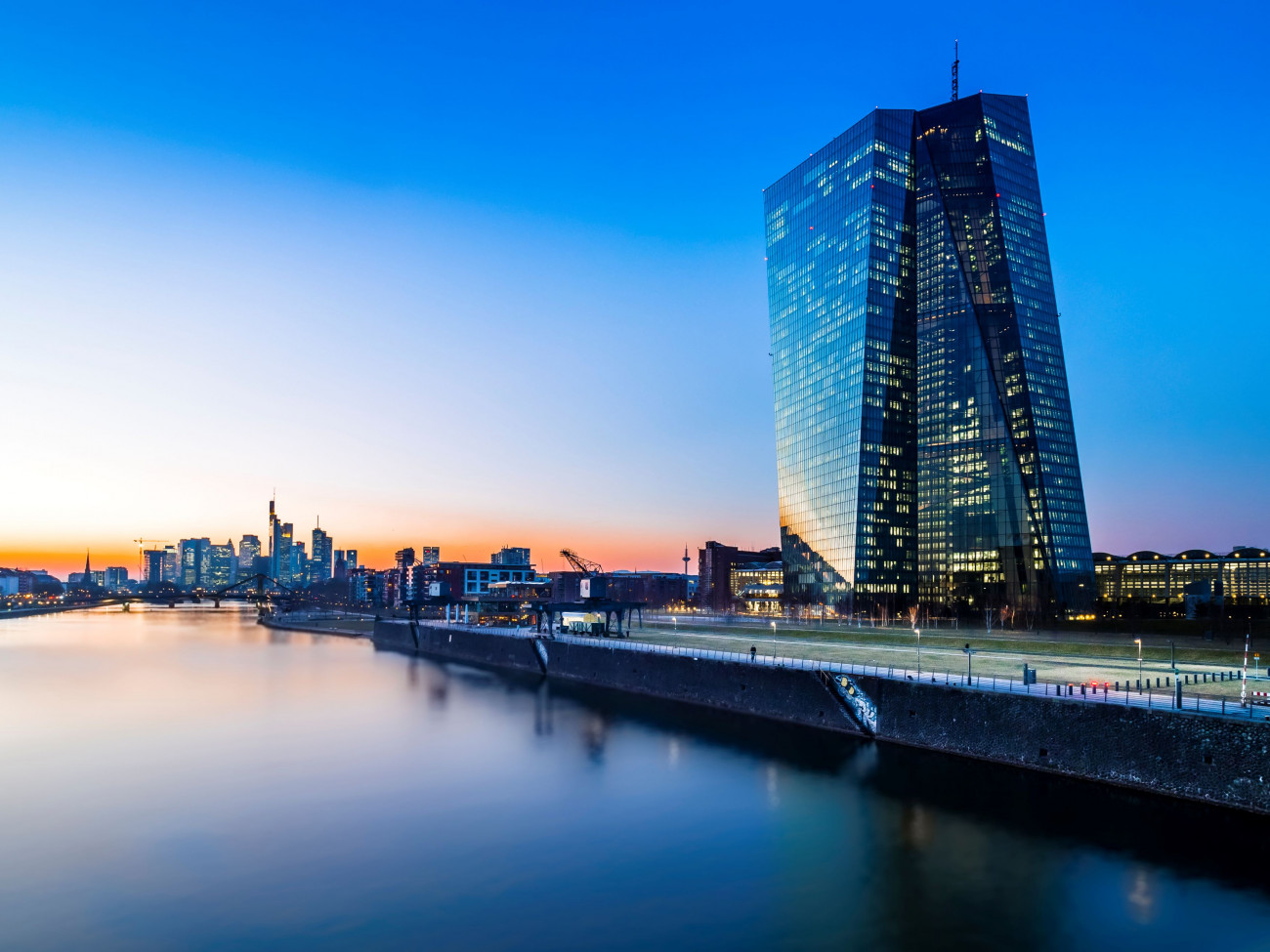 Slika zgradne evropske centralne banke in neboticnika v soncnem zahodu in recni kanal v ospredju 4.2.2024